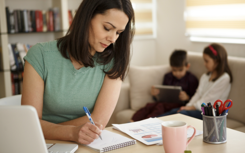 Mother writing things down her to-do list on a piece of paper with her children in the background. This depicts the invisible mental load of motherhood.
