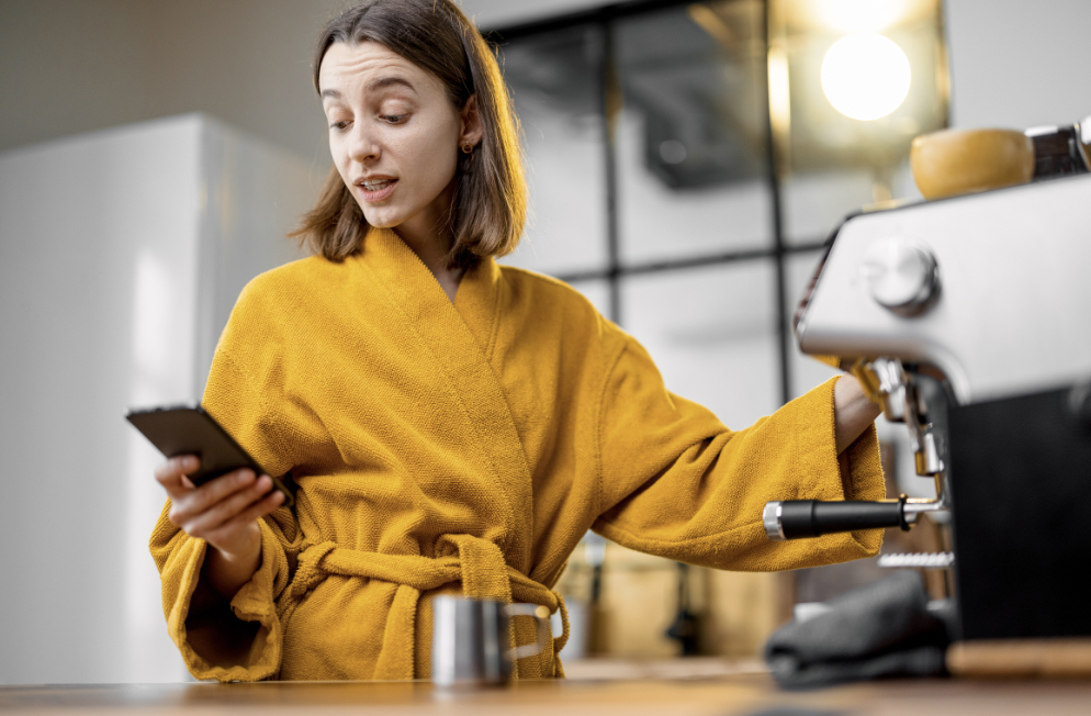 A mother using her phone in the morning as she makes her coffee. She is setting a schedule for when she can be on her phone during the day in order to not spend too much time on her phone when she's around her child(ren).